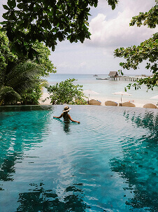 A person relaxes in an infinity pool surrounded by tropical greenery looking out toward a calm ocean and a wooden pier in the distance