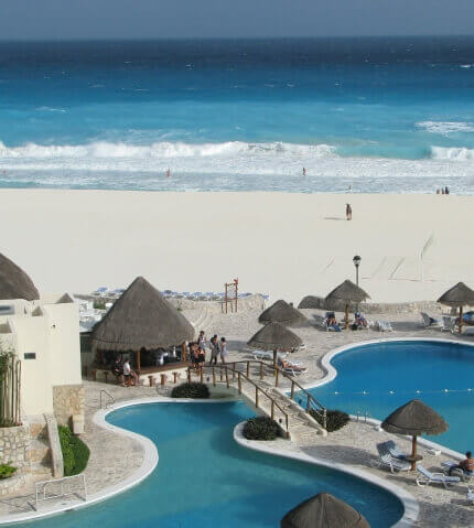 Resort pools with curved shapes and thatched-roof huts overlook a wide sandy beach and turquoise ocean waves in the distance.