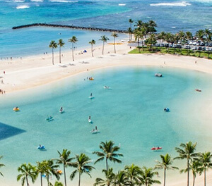 A calm lagoon with people paddleboarding and kayaking is surrounded by palm trees and a sandy beach next to the open ocean.