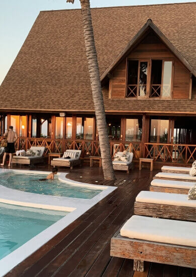 Wooden resort building with a steep roof and a poolside deck lined with cushioned lounge chairs and tropical decor during sunset