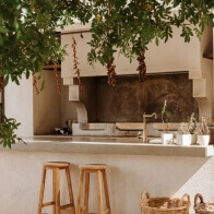 Outdoor kitchen with a rustic design featuring a countertop wooden stools potted plants and hanging dried peppers under leafy tree shade